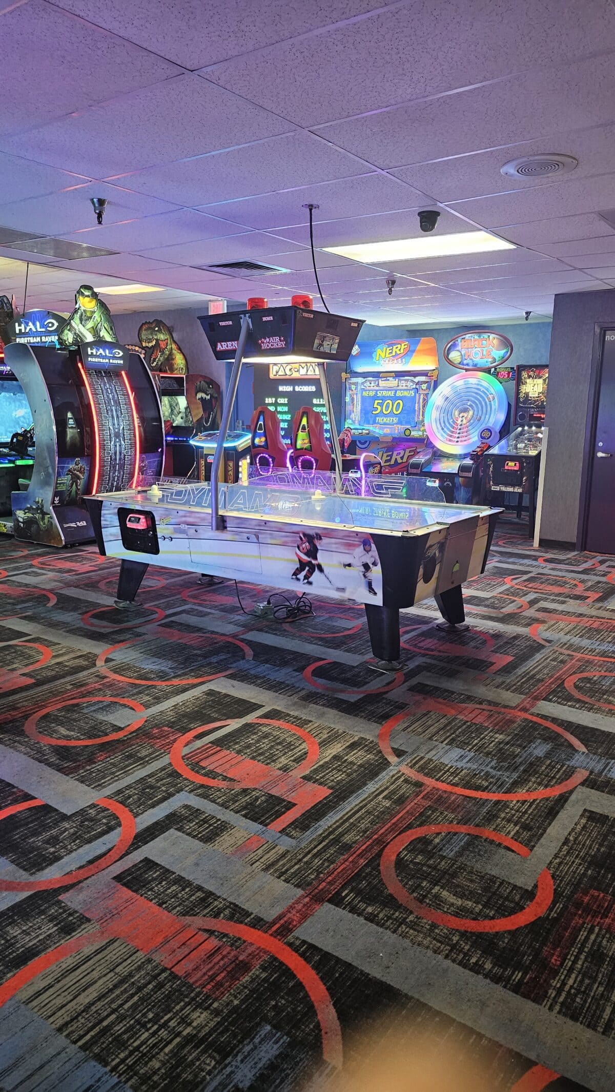 An air hockey table is set up in an arcade with various games and machines, including racing and shooting games, visible in the background.