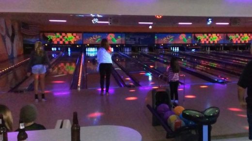People bowling in a dimly lit alley with colorful lights. Various balls and pins are visible, along with spectators in the foreground.