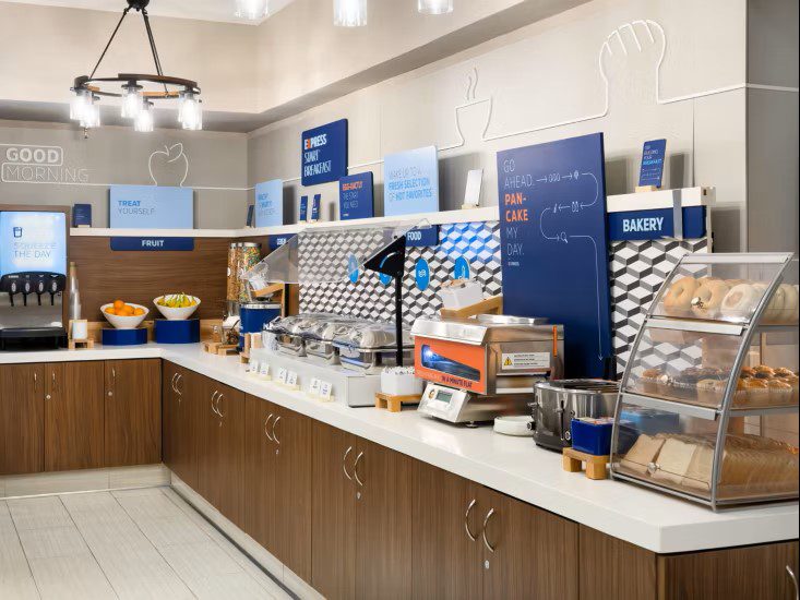 Hotel breakfast buffet area with cereal dispensers, fruit, hot food trays, bagels, bread, a pancake maker, and various signs above the counter in a modern setting.