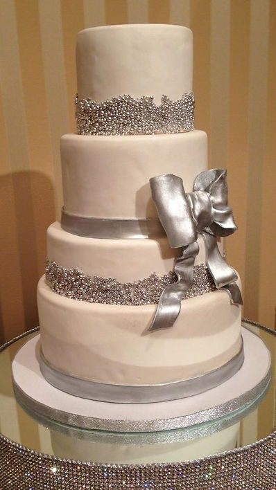 Four-tier white cake with silver ribbon and bow, adorned with silver bead decorations, placed on a mirrored cake stand.