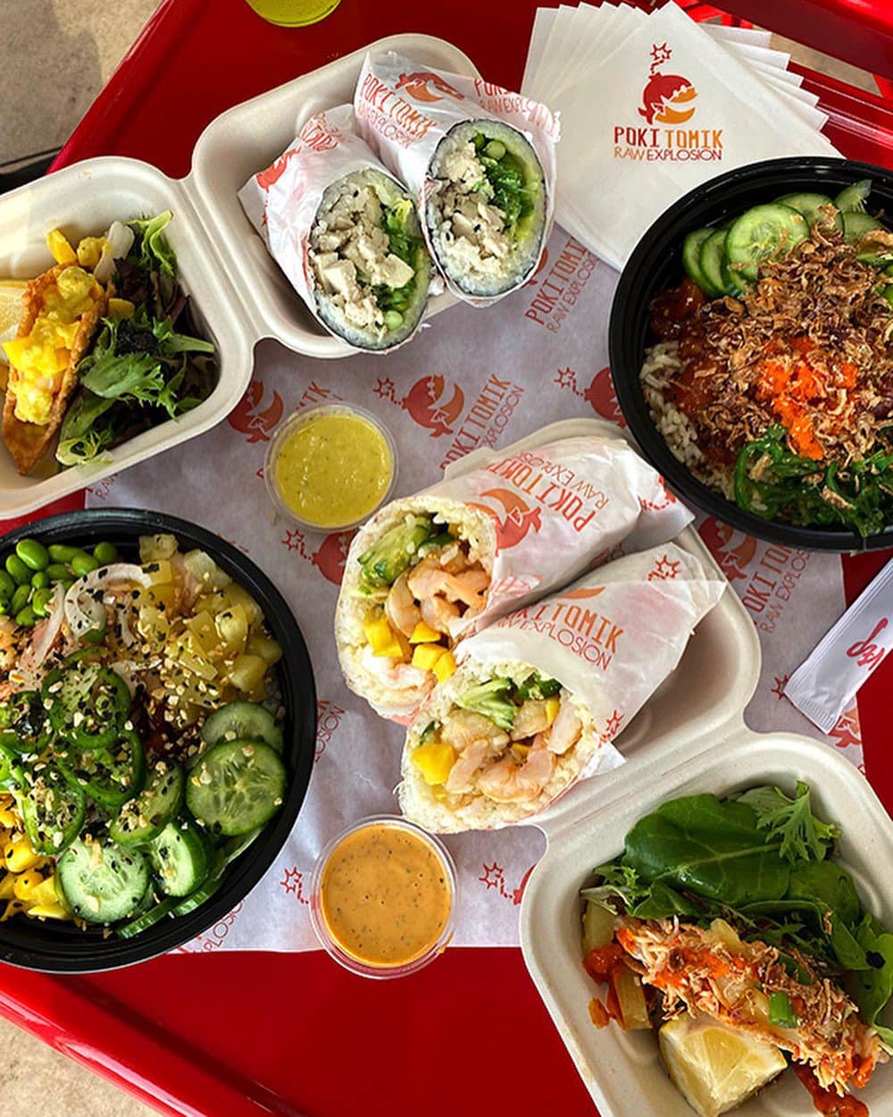 Assorted dishes including poke bowls and sushi burritos with various toppings on a red table, accompanied by sauces in small containers.
