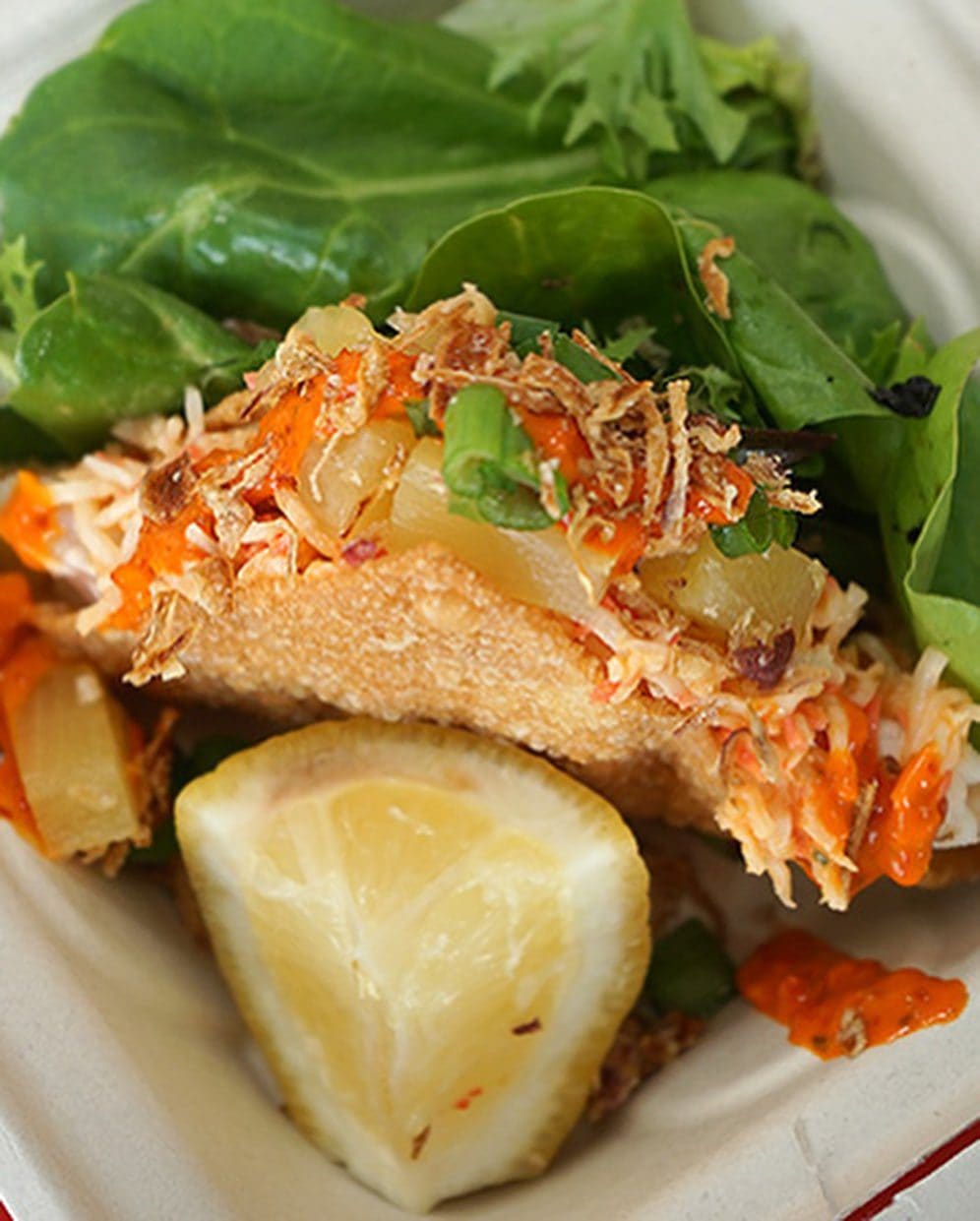 A crispy taco shell filled with shredded vegetables, topped with sauce and herbs, served with fresh greens and a lemon wedge.