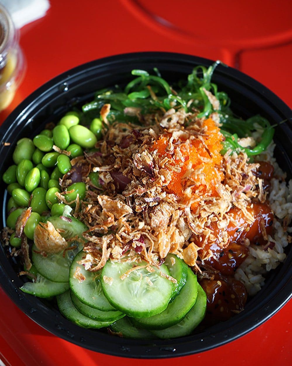 A poke bowl with sliced cucumbers, edamame, seaweed, fried onions, and a central orange topping on rice, served in a black bowl on a red surface.