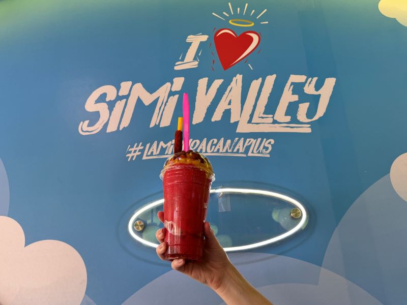 A hand holds a red beverage topped with fruit and spice, against a blue wall with "I ❤️ Simi Valley" and "#IAMLOGANAPLUH" text.
