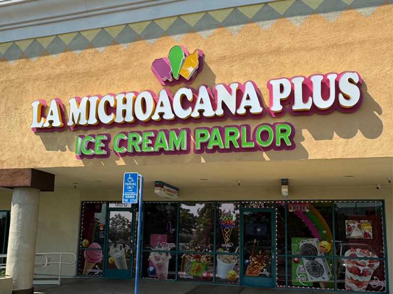 Storefront of La Michoacana Plus Ice Cream Parlor with images of ice cream and treats on the windows. A wheelchair-accessible parking sign is in front.