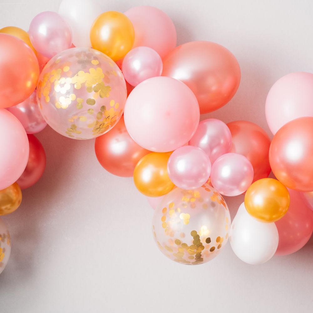A cluster of pink, orange, and white balloons with some featuring gold confetti inside, arranged against a light background.