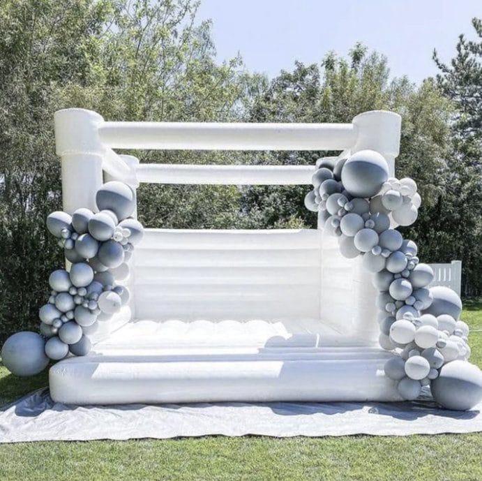 White inflatable bounce house decorated with clusters of gray and white balloons, set on grass with trees in the background.