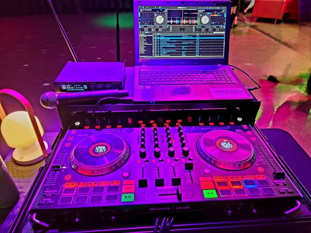 DJ setup with a laptop displaying mixing software, a digital controller with colorful buttons and dials, a microphone, and ambient lighting.