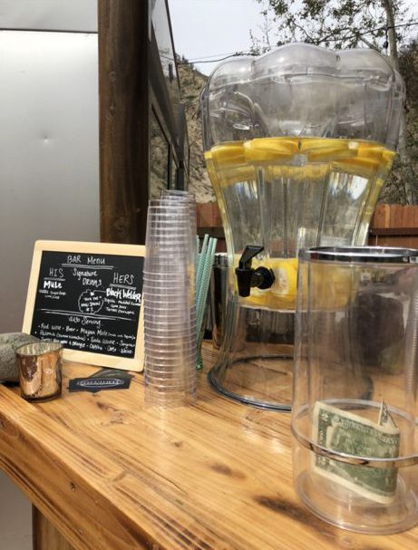 A wooden bar setup with a large water dispenser containing lemon slices, stacked clear cups, a chalkboard menu, and a tip jar with cash on a table.