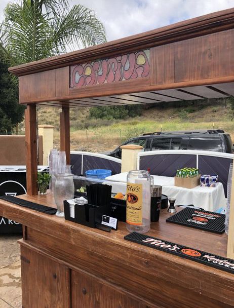 Outdoor bar setup with wooden counter, liquor bottles, plastic cups, and cocktail accessories. A car and greenery are visible in the background.