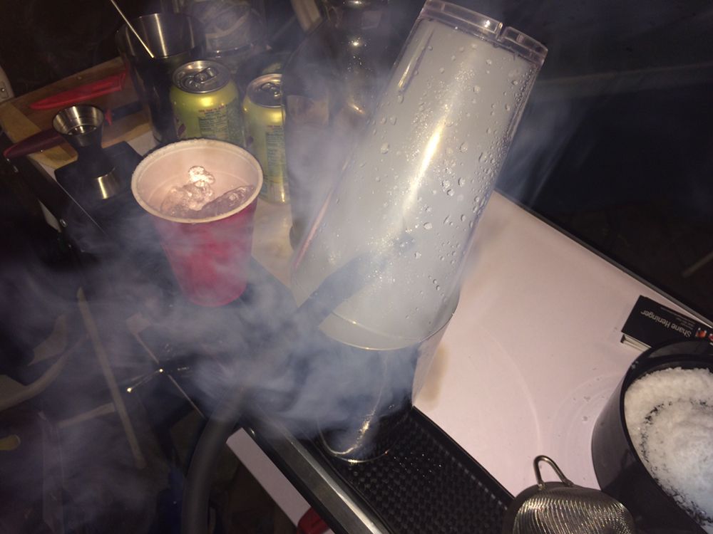 A cup of ice, cans, and a device emitting smoke on a cluttered bar counter.