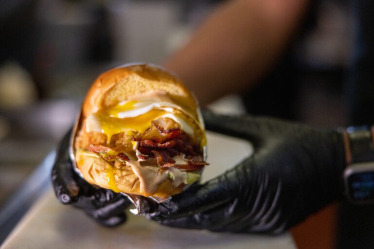 A person wearing black gloves holds a hamburger with bacon, melted cheese, onions, and sauce on a bun.