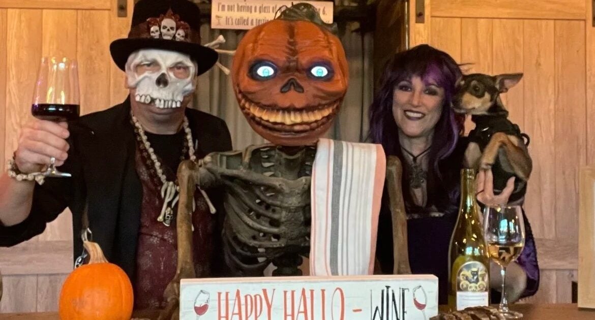 Two adults in Halloween costumes pose with a pumpkin-headed skeleton, a dog, wine glasses, and a “Happy Hallo-Wine” sign on a wooden bar with a pumpkin.