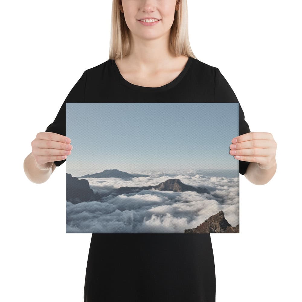 Una persona con una camisa negra sostiene una impresionante estampa titulada "Lienzo mar de nubes desde el Roque de los Muchachos", creada por Volcanica Productions. La imagen captura nubes que envuelven picos de montañas bajo un cielo azul claro, que recuerda la serena belleza de La Palma.