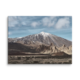 Una montaña nevada bajo un cielo azul nublado, acompañada de formaciones rocosas escarpadas y vegetación escasa en primer plano, captura la belleza natural de La Palma. Esta pintoresca escena se puede transformar en el 