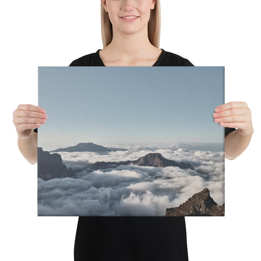 Una persona vestida con una camisa negra y sonriente sostiene un lienzo impreso titulado "Lienzo mar de nubes desde el Roque de los Muchachos", que representa una vista panorámica de las majestuosas montañas de La Palma con sus picos atravesando un mar de nubes bajo el cielo azul claro, que recuerda a Paisajes Canarios.