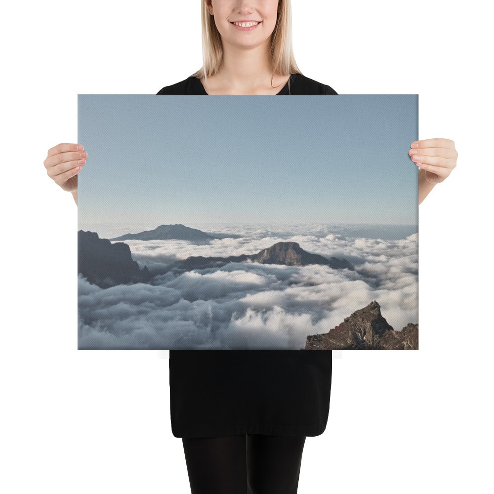 Una persona vestida de negro sostiene el impresionante Lienzo mar de nubes desde el Roque de los Muchachos, que muestra un paisaje montañoso con picos que se elevan majestuosamente sobre un mar de nubes bajo el cielo azul claro, capturando la belleza etérea de La Palma.