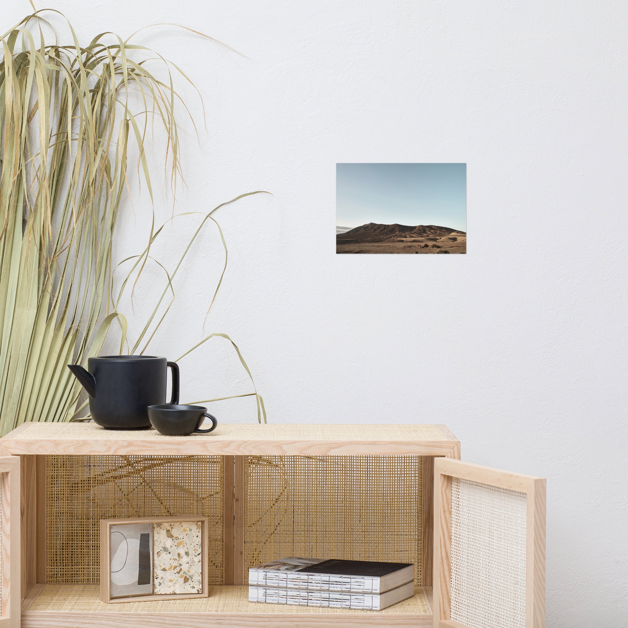 Una habitación minimalista cuenta con un armario de madera clara que exhibe una tetera y una taza negras. Arriba, un póster de Los Ajaches está colgado en la pared blanca. Hierba seca alta adorna el lado izquierdo, mientras que varios libros de Volcanica Productions descansan cerca del armario.