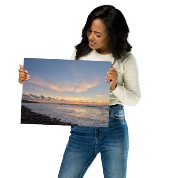 Una mujer con una camisa blanca de manga larga y jeans sostiene una gran fotografía de un paisaje costero al atardecer, titulada "Póster atardecer en Los Guirres" de Volcanica Productions. Esta pieza captura una costa de guijarros, un mar en calma y nubes que reflejan tonos cálidos. La mujer examina el cartel de cerca, apreciando cada detalle.