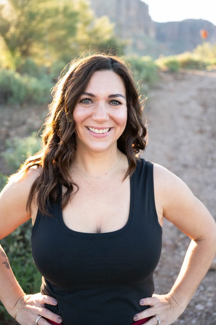 A woman with long brown hair wearing a sleeveless black top stands outdoors on a sunlit, rocky path, smiling at the camera with her hands on her hips.