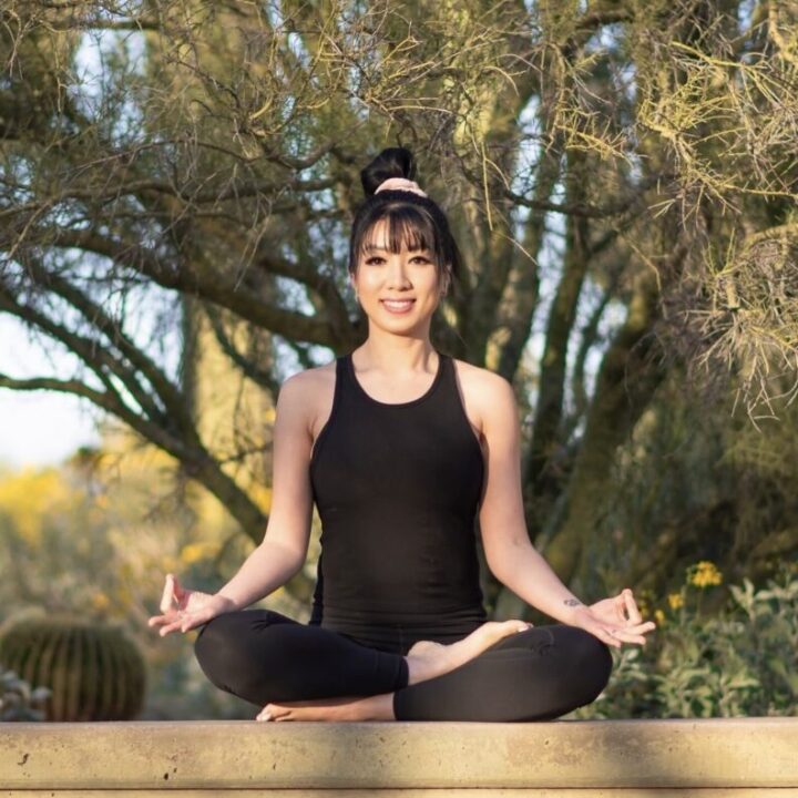 Person sitting cross-legged in a yoga pose outdoors, wearing a black outfit. Trees and greenery are visible in the background.