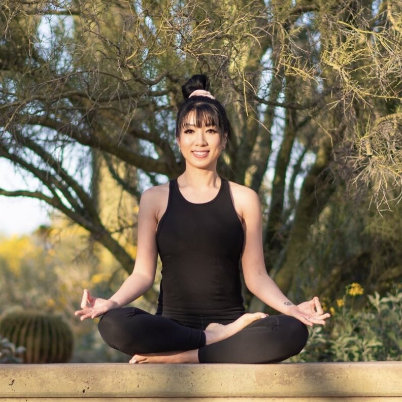 Person sitting cross-legged in a yoga pose outdoors, wearing a black outfit. Trees and greenery are visible in the background.