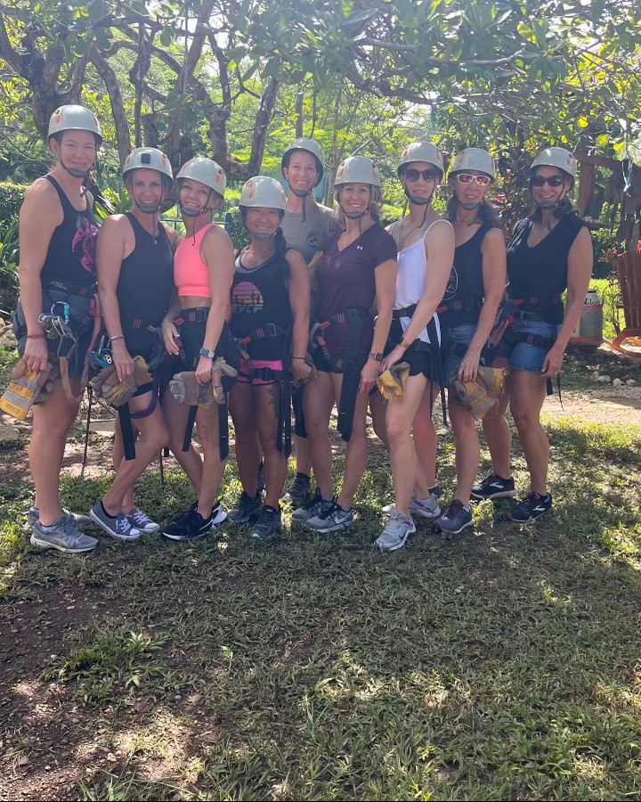 A group of nine people wearing helmets and harnesses stand together outdoors, ready for a zip-lining activity. Trees and grass are visible in the background.