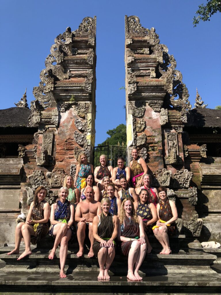 A group of people in colorful attire sit on steps in front of an ornate stone temple gate under a clear blue sky.