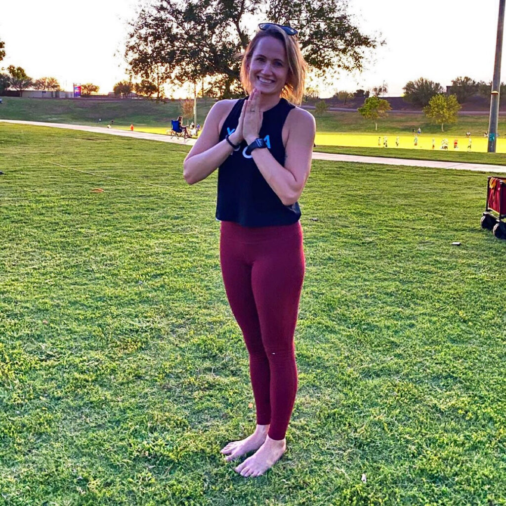 A woman stands barefoot on grass with hands together in a greeting gesture, wearing a black top and red leggings, with trees and people in the background.