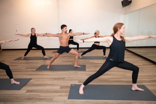 A group of people in a yoga studio are practicing Warrior II pose on mats, facing a mirrored wall.
