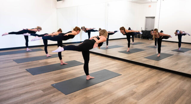 A group of people practice standing yoga poses in a mirrored studio, balancing on one leg with arms extended back, on individual mats.