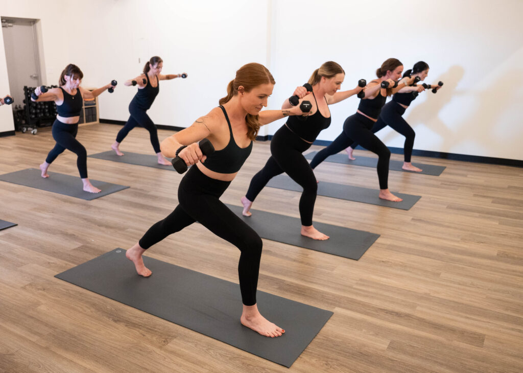 A group of people in exercise attire perform a lunge with dumbbells on yoga mats in a studio.