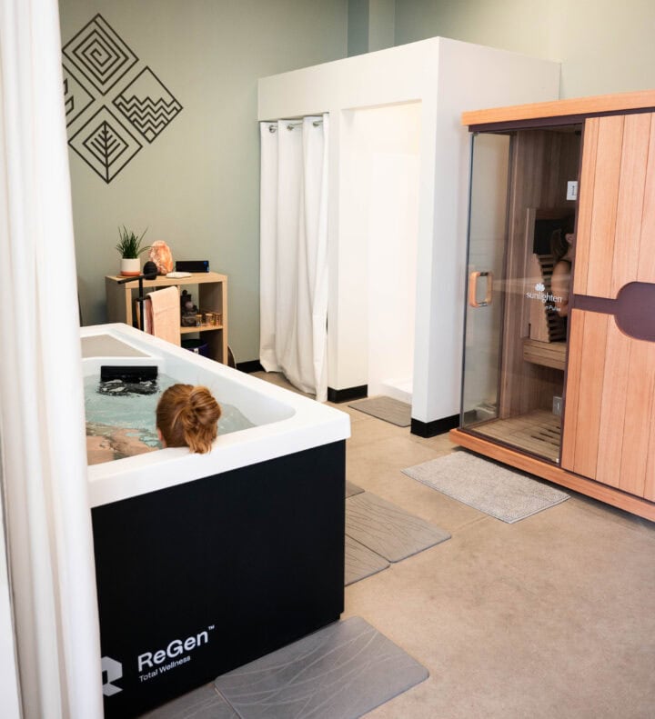 A woman relaxes in a ReGen bath next to a wooden sauna in a wellness clinic room with modern decor.