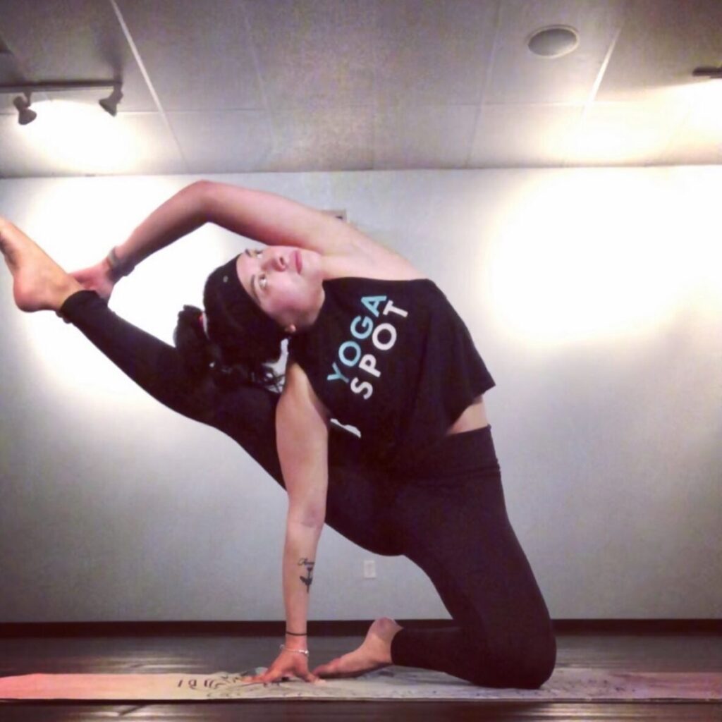 Person in a yoga pose, wearing a black tank top and leggings, holding one leg up with both hands on a yoga mat indoors.