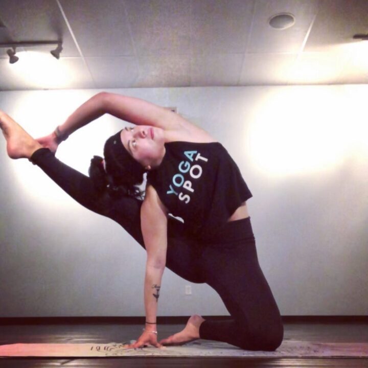 Person in a yoga pose, wearing a black tank top and leggings, holding one leg up with both hands on a yoga mat indoors.