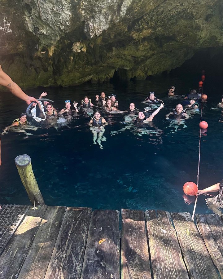 A group of people swim and wave in a large cave pool, surrounded by rock formations. A wooden platform is visible in the foreground.