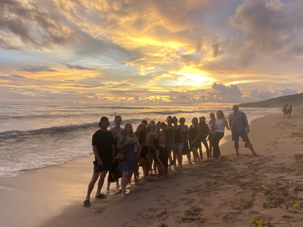 A group of people stands on a beach at sunset, with the ocean and a colorful sky in the background.