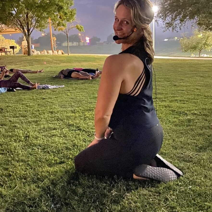 A woman kneels on grass wearing a headset microphone, facing the camera. Other people are lying on the grass in the background.