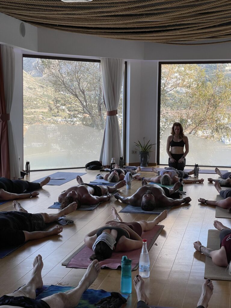 People lying on yoga mats in a spacious room with large windows. An instructor stands at the front. Yoga mats and water bottles are visible.