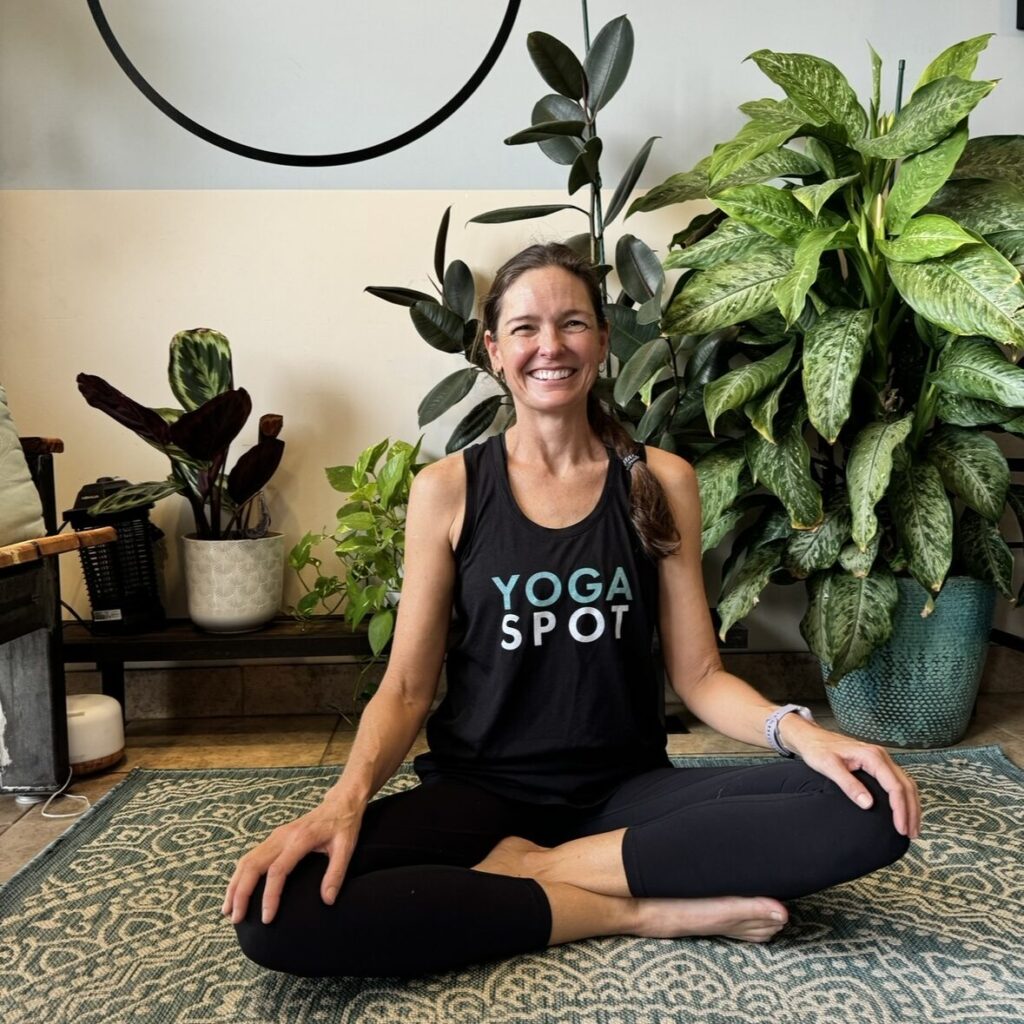 Person sitting cross-legged on a yoga mat, smiling, in front of several potted plants. Wearing a "Yoga Spot" tank top.