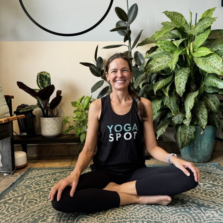 Person sitting cross-legged on a yoga mat, smiling, in front of several potted plants. Wearing a "Yoga Spot" tank top.