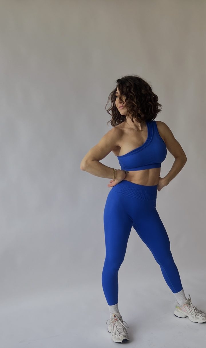 A person wearing a blue one-shoulder workout outfit and white sneakers stands in a confident pose against a plain light background.