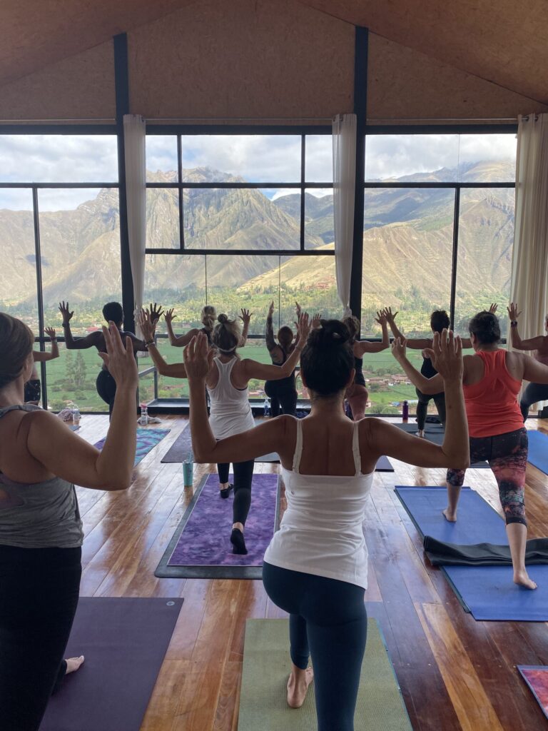 A group of people practice yoga in a mountain-view studio, performing a balance pose on mats with arms raised, facing large windows.