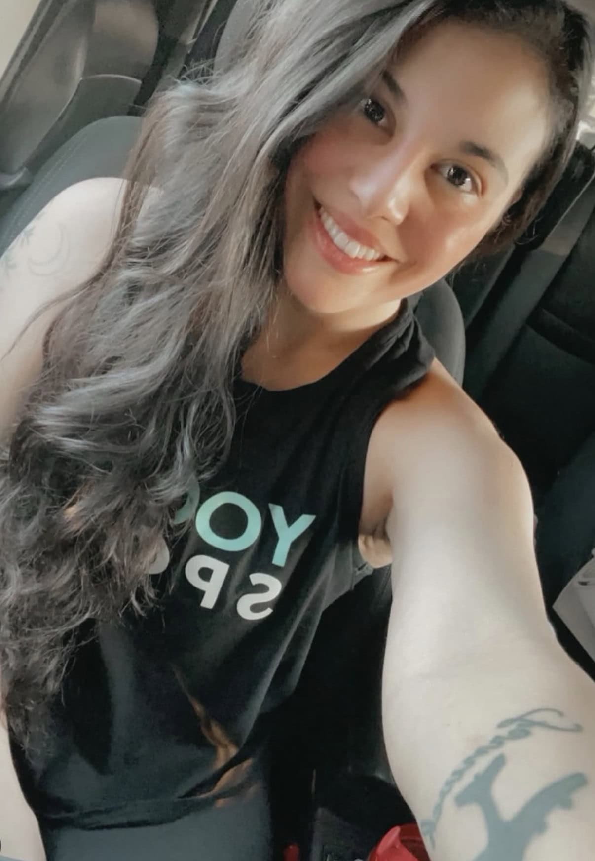 Woman with long dark hair and a tattoo on her arm takes a selfie in a car, smiling and wearing a sleeveless black shirt with partially visible text.
