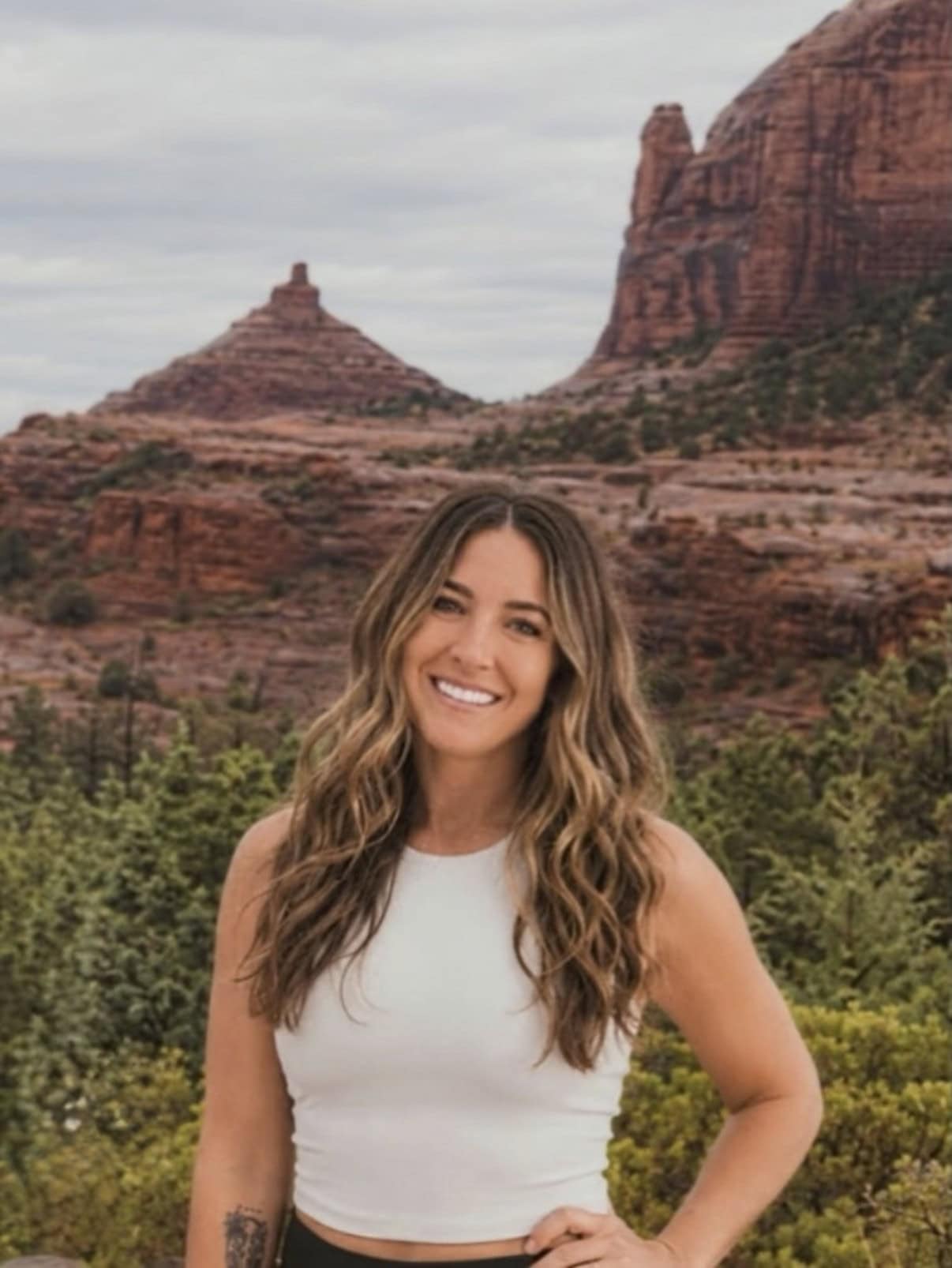 Woman with long wavy hair wearing a white sleeveless top stands smiling in front of red rock formations and green shrubs under a cloudy sky.