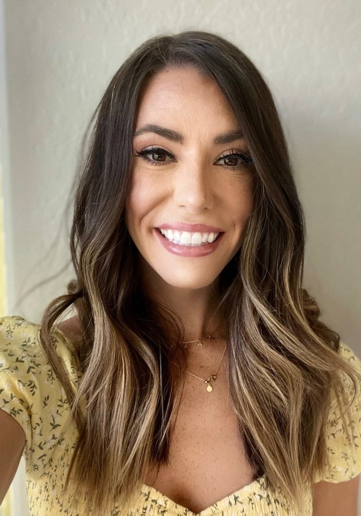 Smiling woman with long wavy brown hair wearing a yellow floral top, standing indoors against a plain light-colored wall.