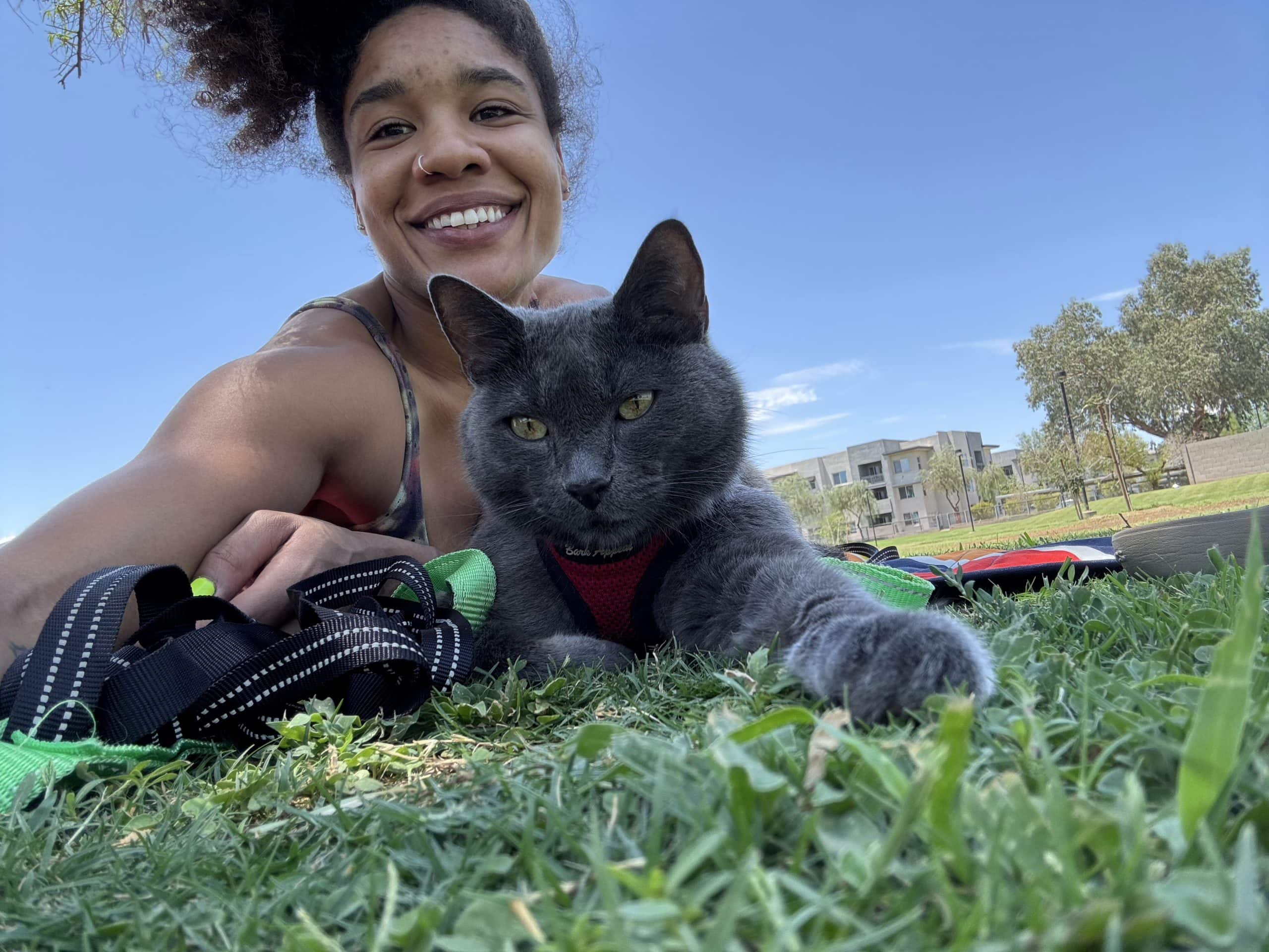 A person lying on grass next to a gray cat wearing a harness. Trees and buildings are visible in the background under a clear blue sky.