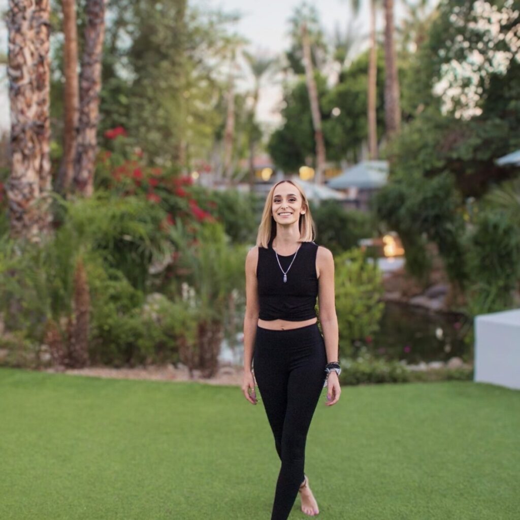 Woman in a black outfit walks barefoot on grass, surrounded by lush greenery and palm trees.