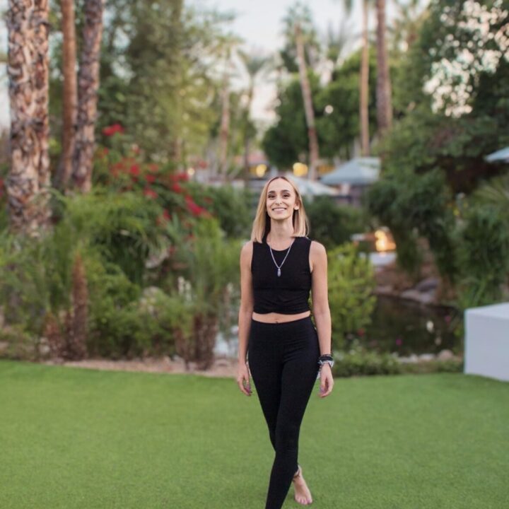 Woman in a black outfit walks barefoot on grass, surrounded by lush greenery and palm trees.