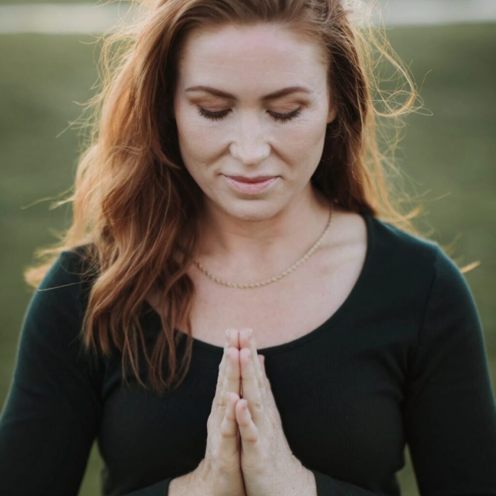 Person with long hair and a black shirt, eyes closed, holding hands in a prayer position outside, with blurred green background.